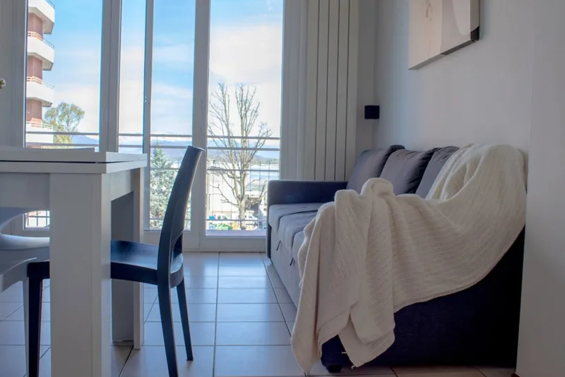 Soggiorno di Casa Martina con divano letto grigio, plaid chiaro e ampia vetrata affacciata sul Lago Maggiore
