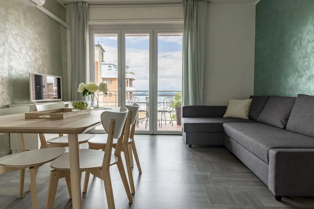 Soggiorno de La Terrazza con divano angolare grigio, tavolo in legno chiaro, pareti verde salvia e portafinestra sul terrazzo con vista lago