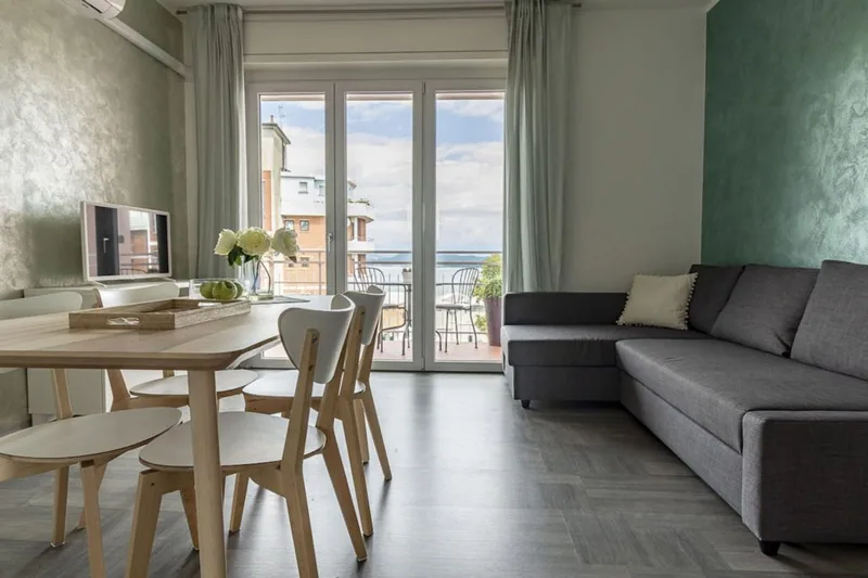 Soggiorno de La Terrazza con divano angolare grigio, tavolo in legno chiaro, pareti verde salvia e portafinestra sul terrazzo con vista lago